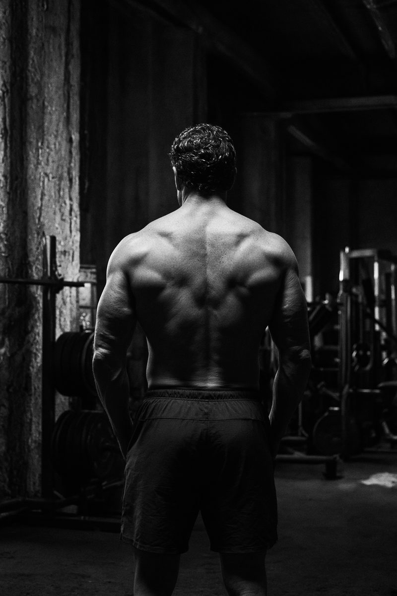 Black-and-white photograph of a developed physique standing in a dim gym, back to camera.
