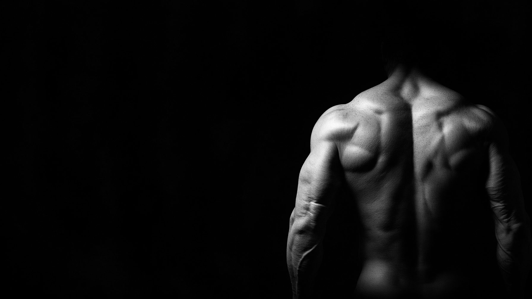 Black-and-white studio photograph of a highly developed physique, back and shoulders against a black field.